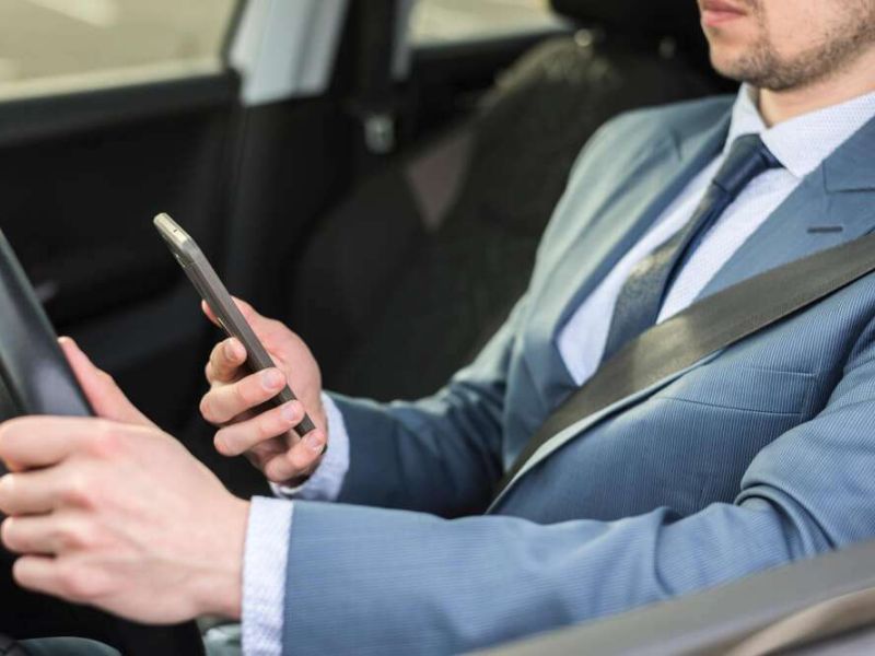 Businessman in a suit driving while holding a smartphone, illustrating distracted driving and safety risks.