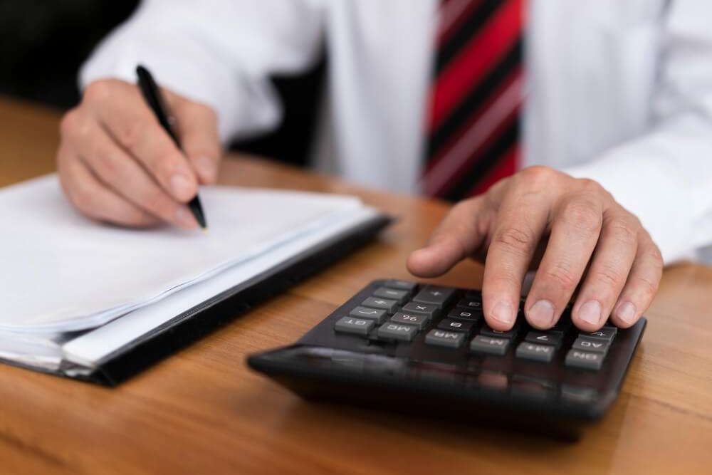 Person using a calculator and writing on paper at a wooden desk, wearing a striped tie and white shirt.