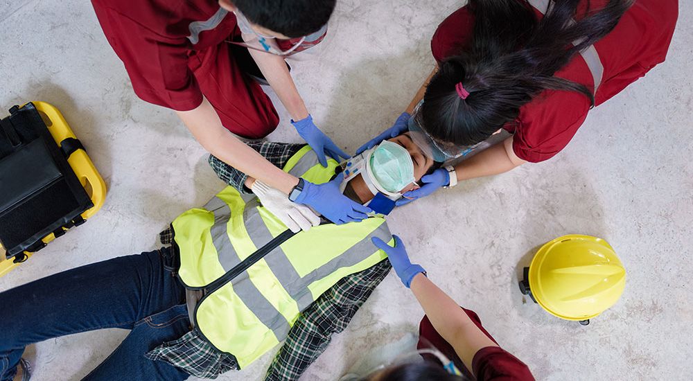 Emergency responders in red uniforms aid an injured worker wearing a reflective vest on the ground, near a hard hat.