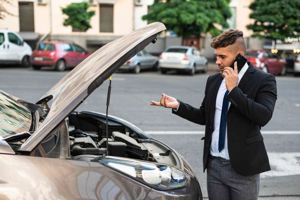 Man in suit on phone with car hood open, possible breakdown in city setting.