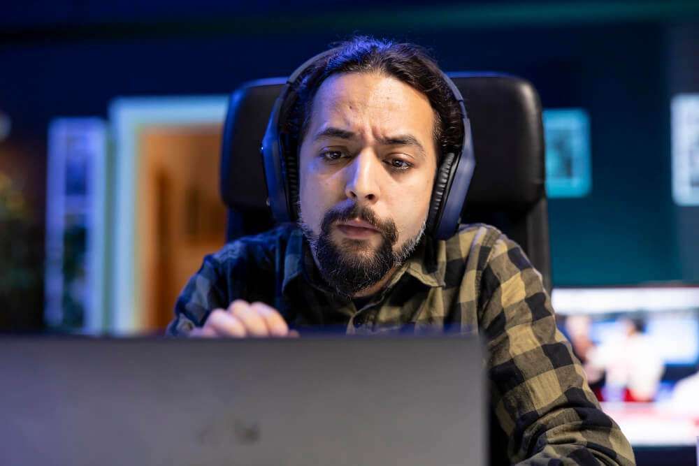 Man with headphones focused on laptop in a dimly lit room.