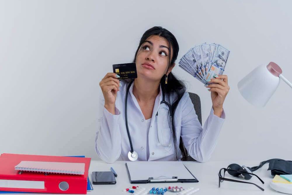Doctor pondering over credit card and cash at desk with medical tools.