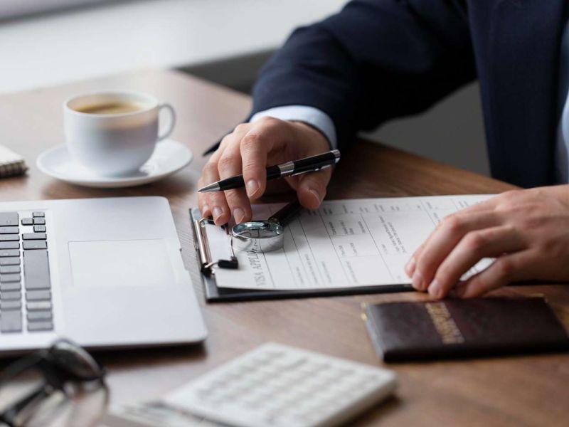 Person filling form on clipboard at desk with laptop and coffee, emphasizing business or office work setting.