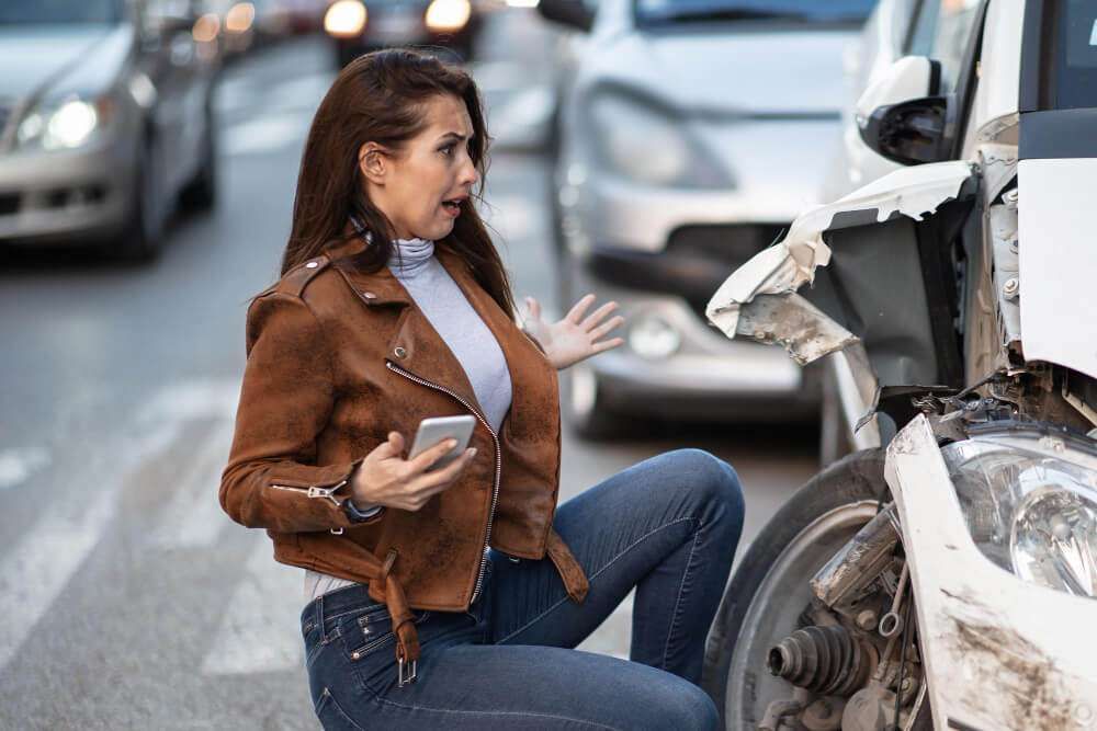 Woman shocked at car accident, holding phone beside damaged vehicle on street.