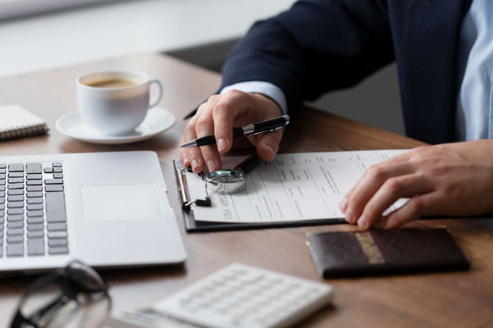 Person filling form on clipboard at desk with laptop and coffee, emphasizing business or office work setting.
