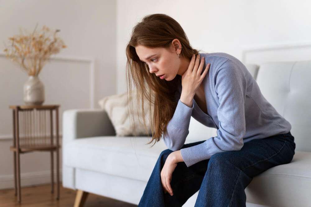 Woman in casual wear sitting on a sofa, looking thoughtful and holding her neck in a cozy living room setting.