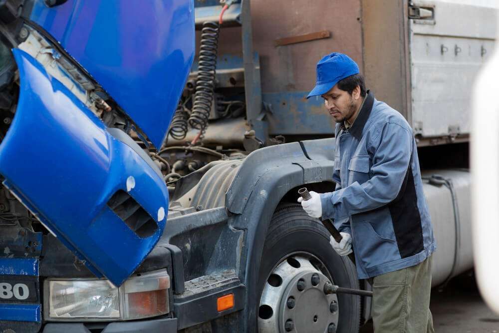 Mechanic in blue cap and jacket repairing a large truck's engine, ensuring maintenance and safety.