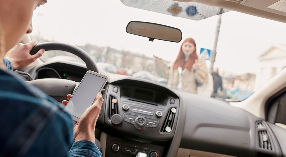 Driver distracted by phone, unaware of pedestrian crossing in front of car, emphasizing road safety.