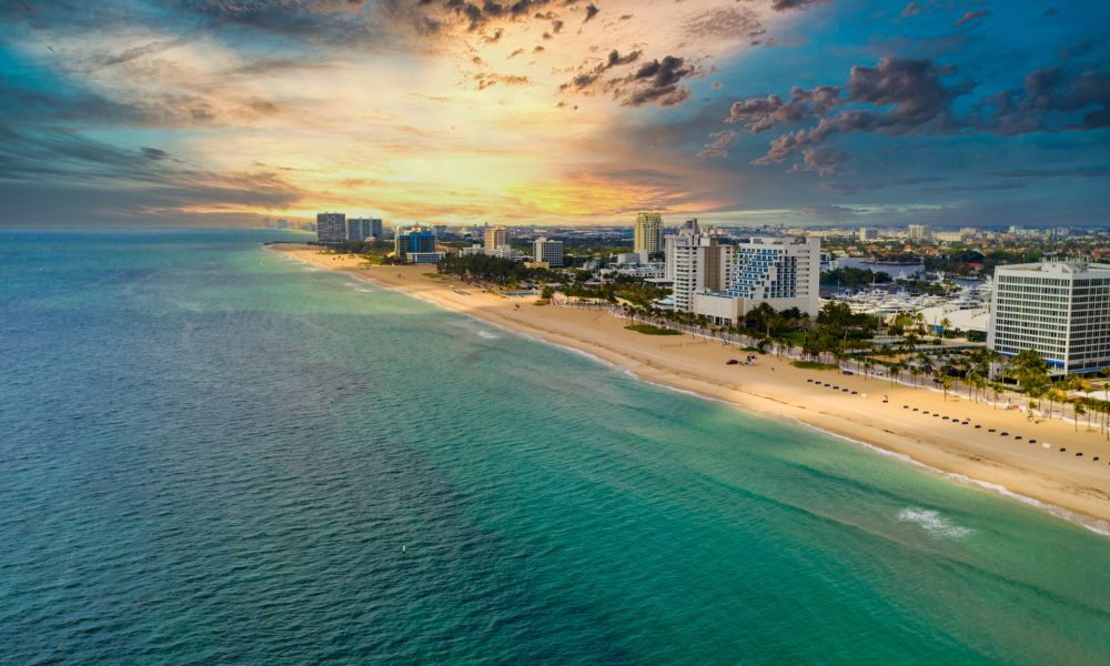 Aerial view of sunny beachfront cityscape at sunset, featuring sandy beaches, turquoise water, and a vibrant skyline.