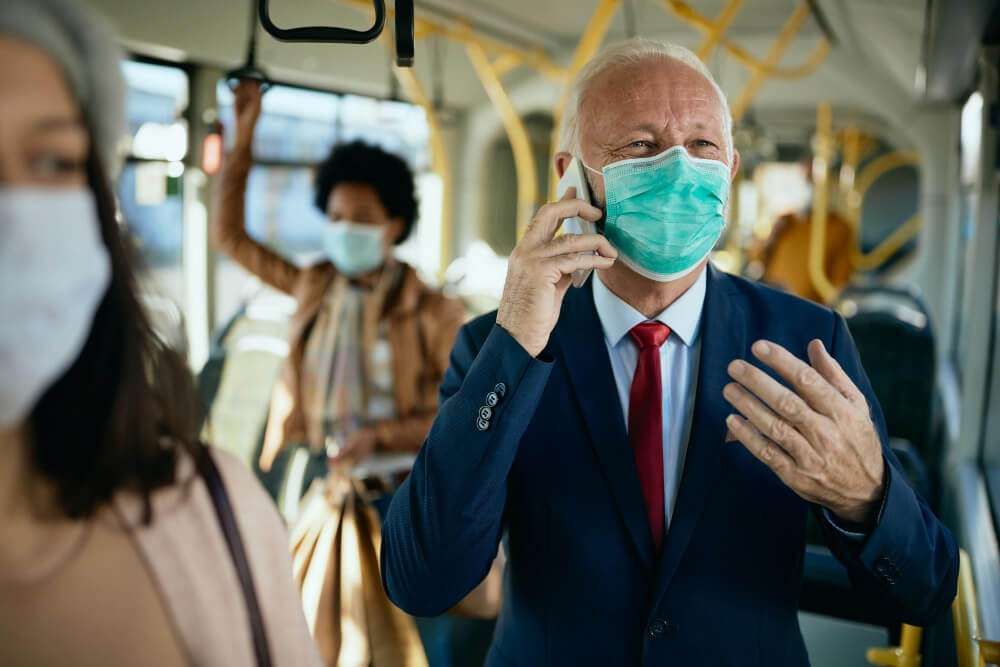 Businessman in a suit and mask talking on the phone while commuting on a bus, with others wearing masks.