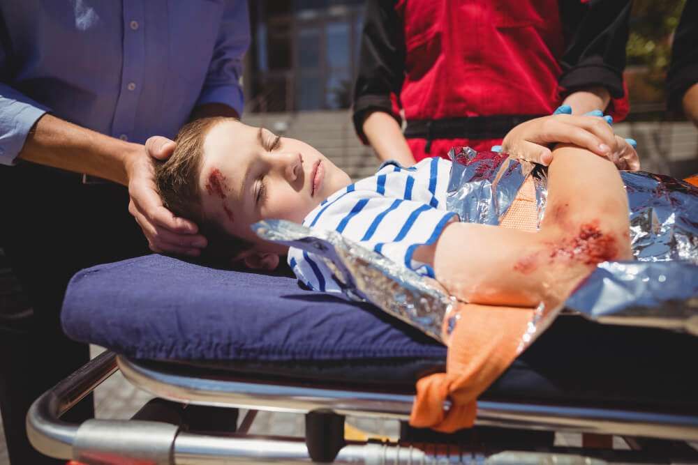 Injured child with head wound on stretcher, receiving first aid assistance outdoors.