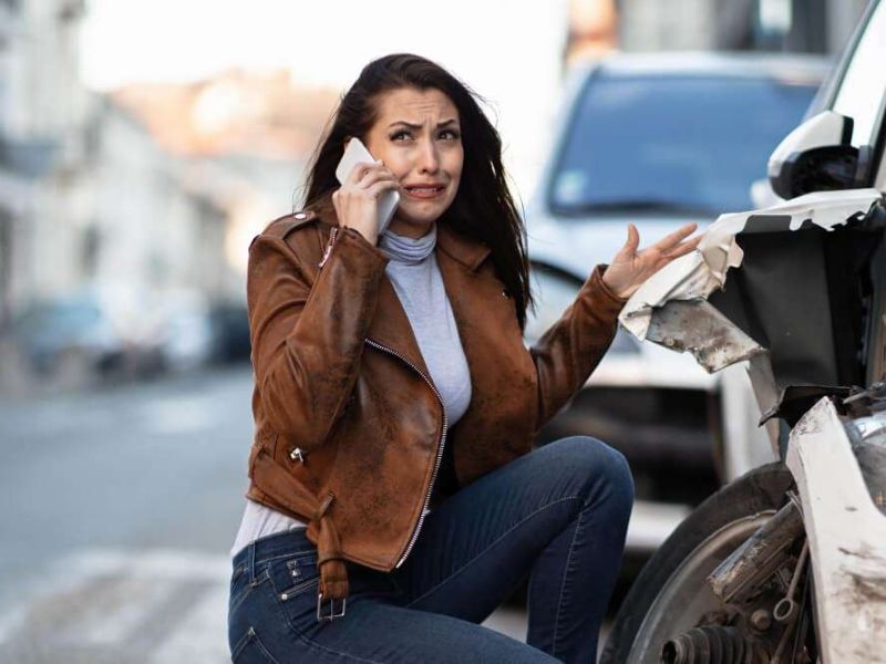 Woman on phone after car accident, inspecting damage with a worried look, on a street with traffic in the background.