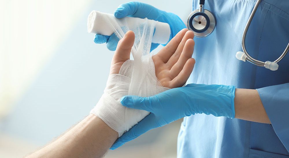 Medical professional bandaging a patient's hand with blue gloves and a stethoscope in a healthcare setting.