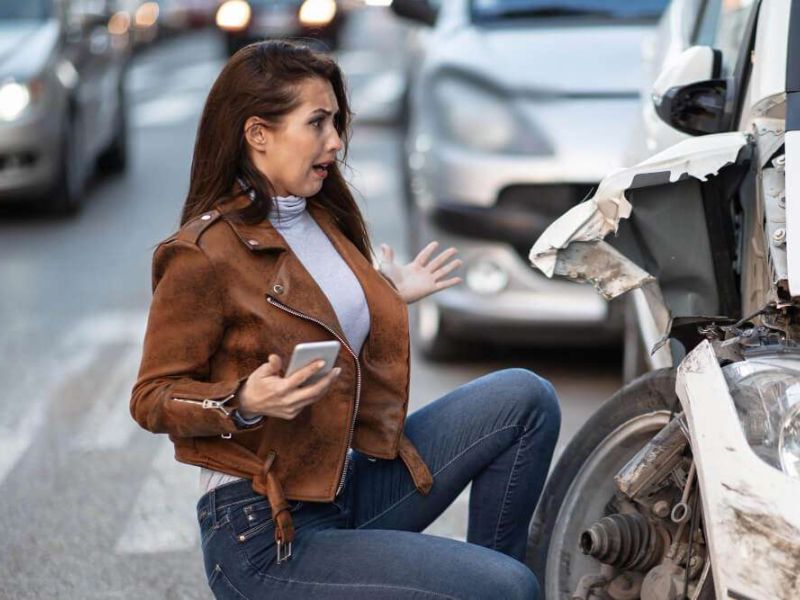 Woman shocked at car accident, holding phone beside damaged vehicle on street.