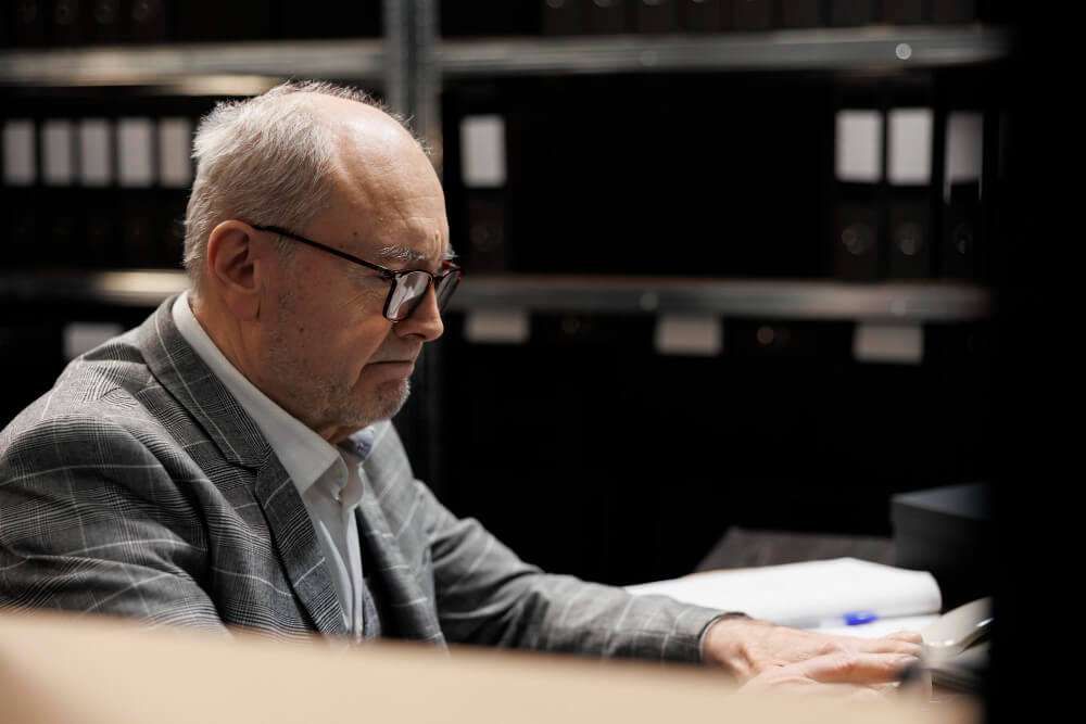 Elderly man in glasses working on a computer in a dimly lit archive room, shelves filled with binders in background.