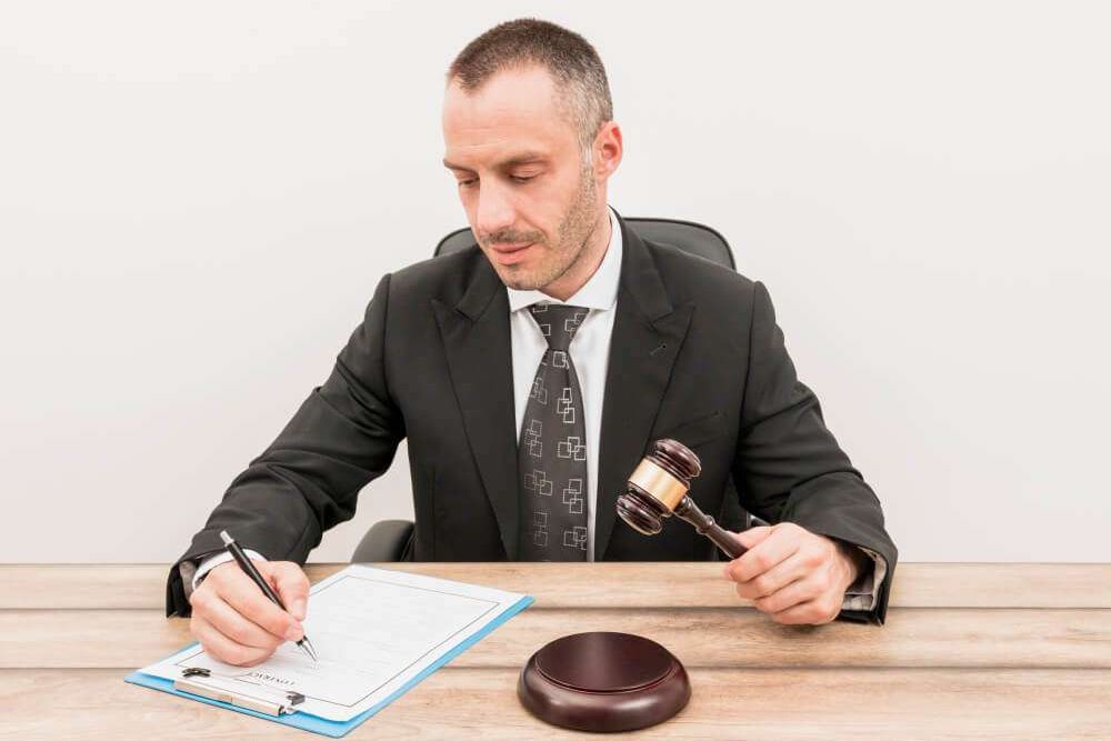 A judge in a suit signing a document while holding a gavel, seated at a wooden desk.