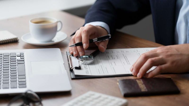 Person filling form on clipboard at desk with laptop and coffee, emphasizing business or office work setting.