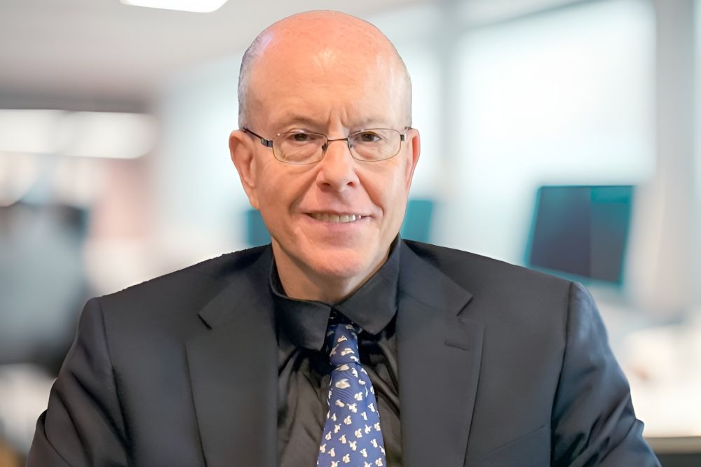 Bald man in dark suit and glasses smiling in an office setting with blurred background.