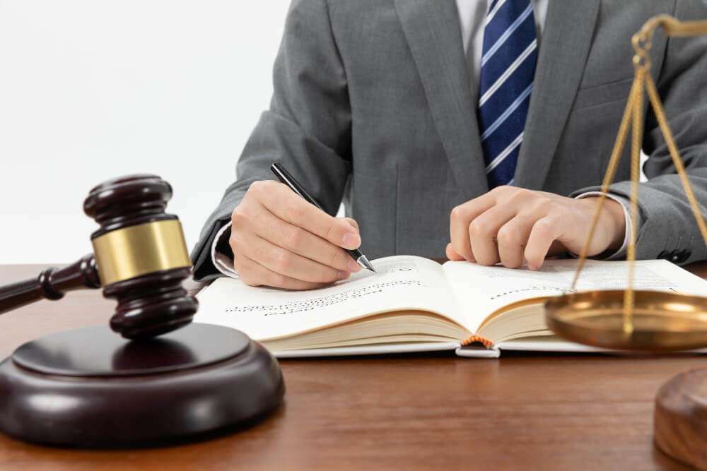 Lawyer writing in book at desk with gavel and scales, representing legal work and justice concept.