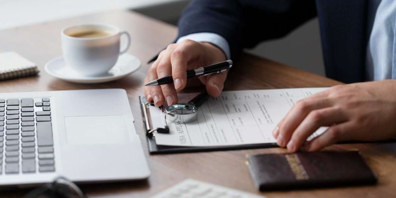 Person filling form on clipboard at desk with laptop and coffee, emphasizing business or office work setting.