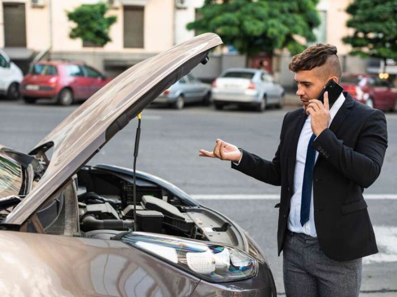 Man in suit on phone with car hood open, possible breakdown in city setting.