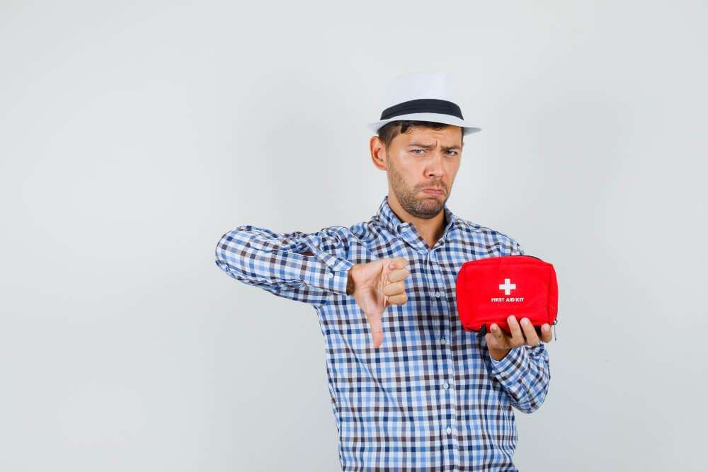 Man in hat giving thumbs down, holding a red first aid kit against a gray background.