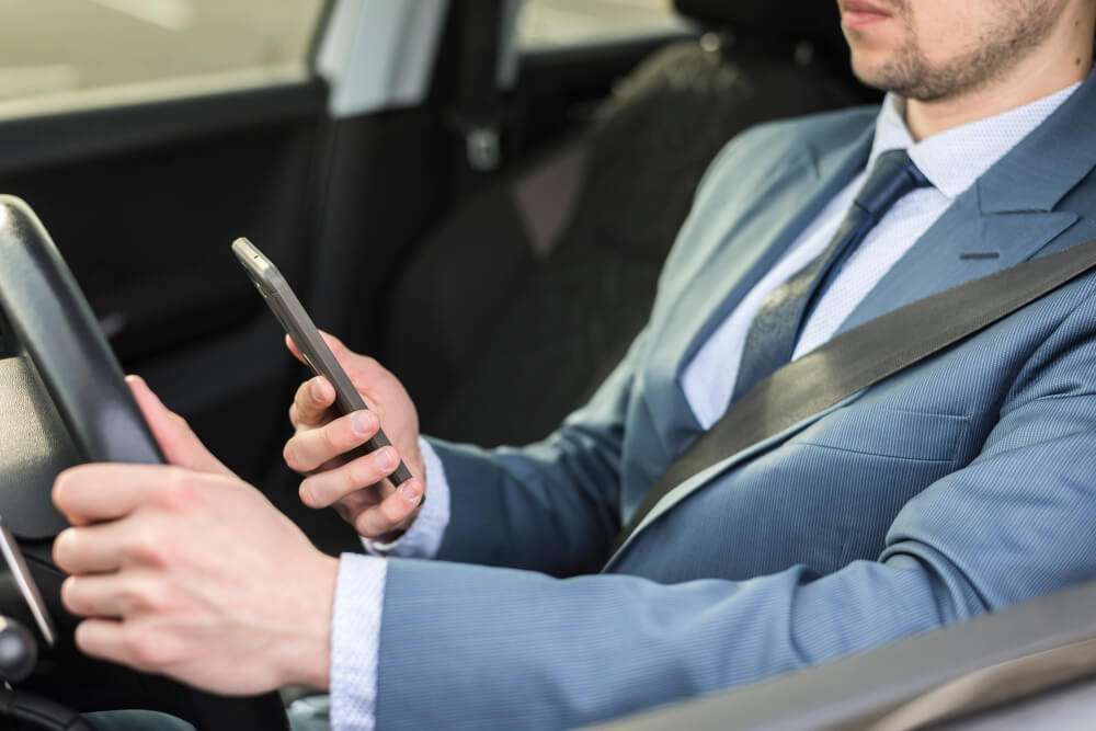 Businessman in a suit driving while holding a smartphone, illustrating distracted driving and safety risks.