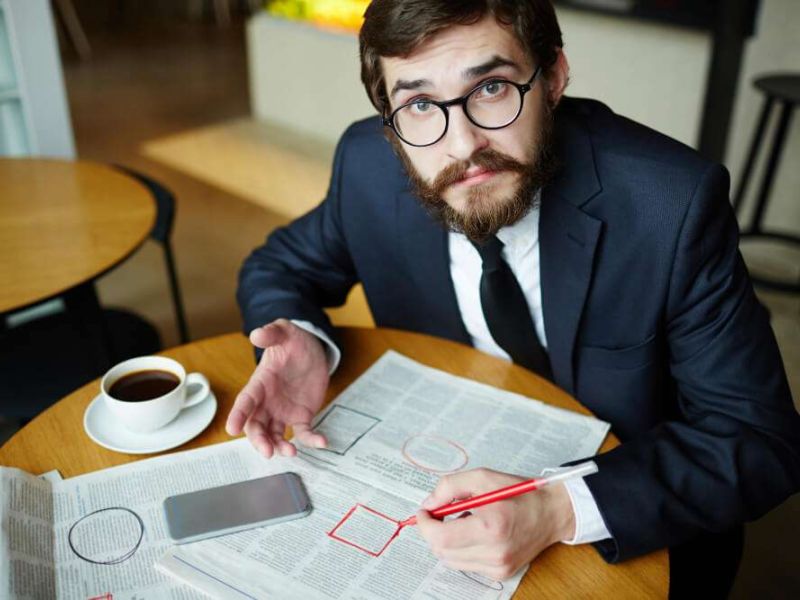 Man in suit reading newspaper at cafe table, circling classified ads with a pen, coffee and smartphone nearby.