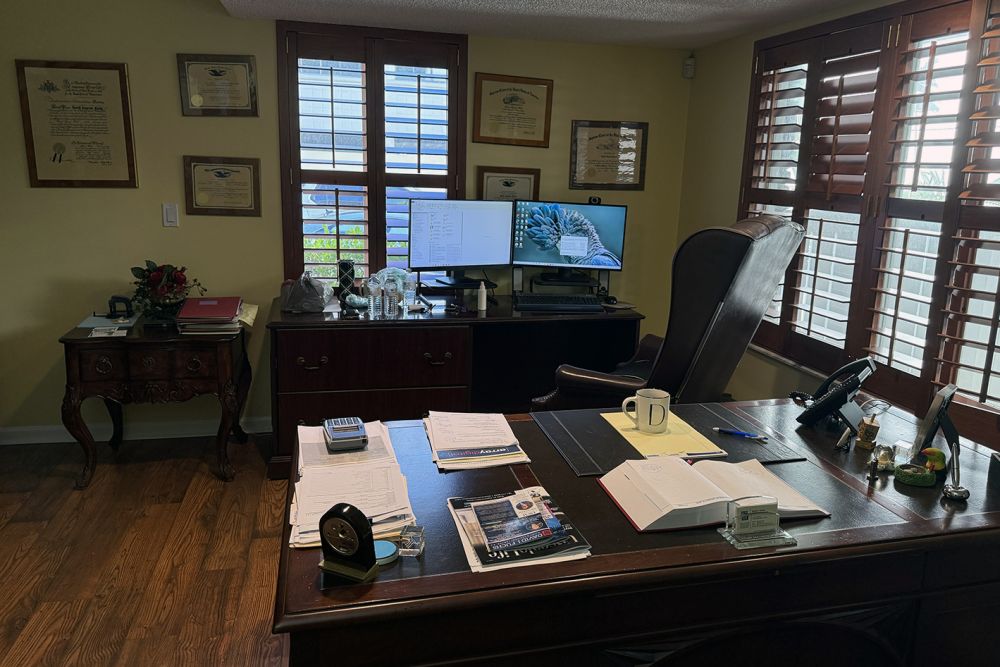 Home office setup with dual monitors, wooden desk, certificates on the wall, and large windows with wooden shutters.