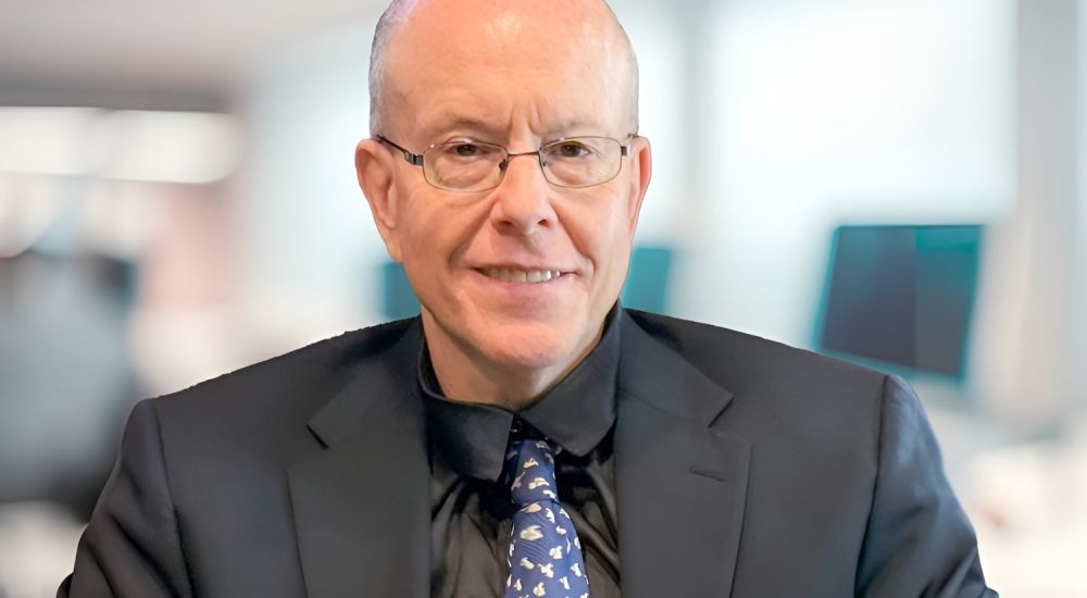 Bald man in dark suit and glasses smiling in an office setting with blurred background.