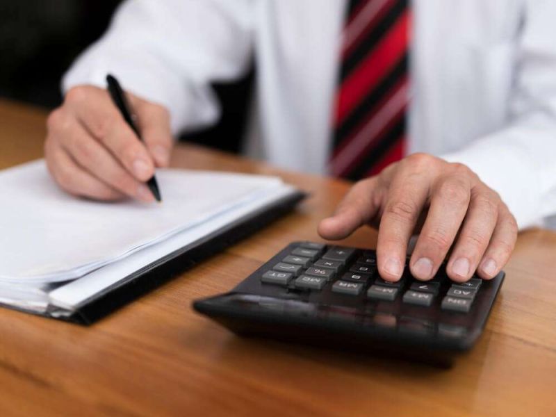 Person using a calculator and writing on paper at a wooden desk, wearing a striped tie and white shirt.