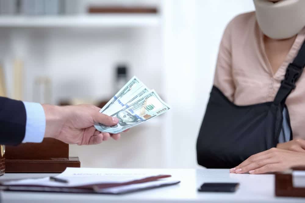 Hand giving cash to person with arm sling and neck brace, symbolizing personal injury compensation or settlement.