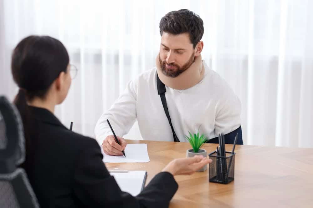 Man with arm sling and neck brace signs documents in office meeting.