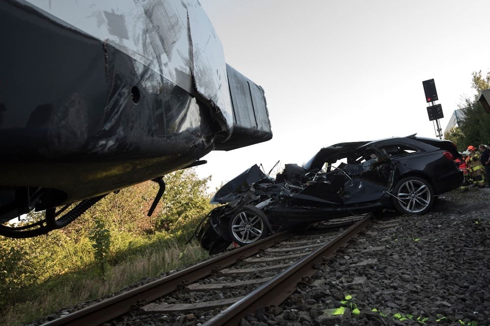 Train collision with car on tracks, severe damage visible, emergency responders in background, railroad safety issue.