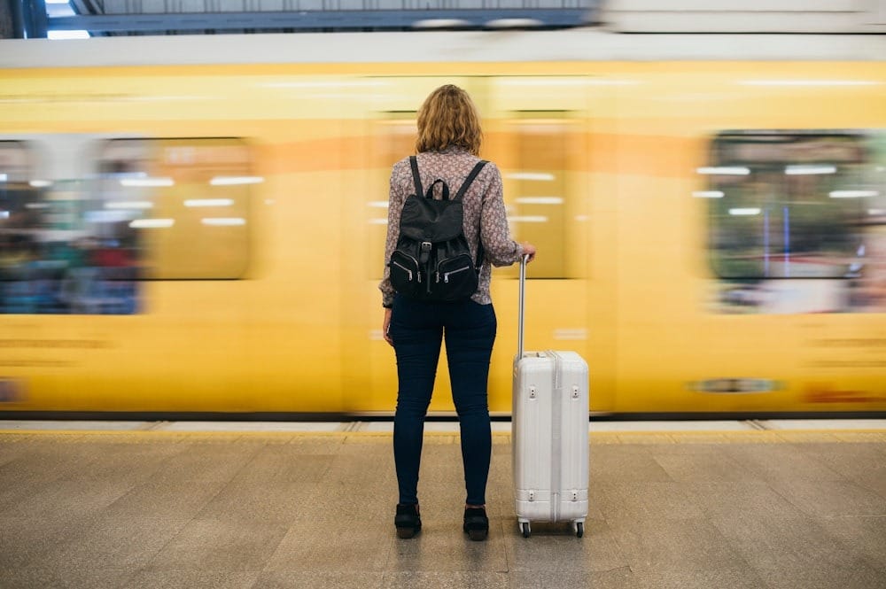 Traveler with suitcase and backpack waits as yellow train rushes by, capturing journey anticipation at station.