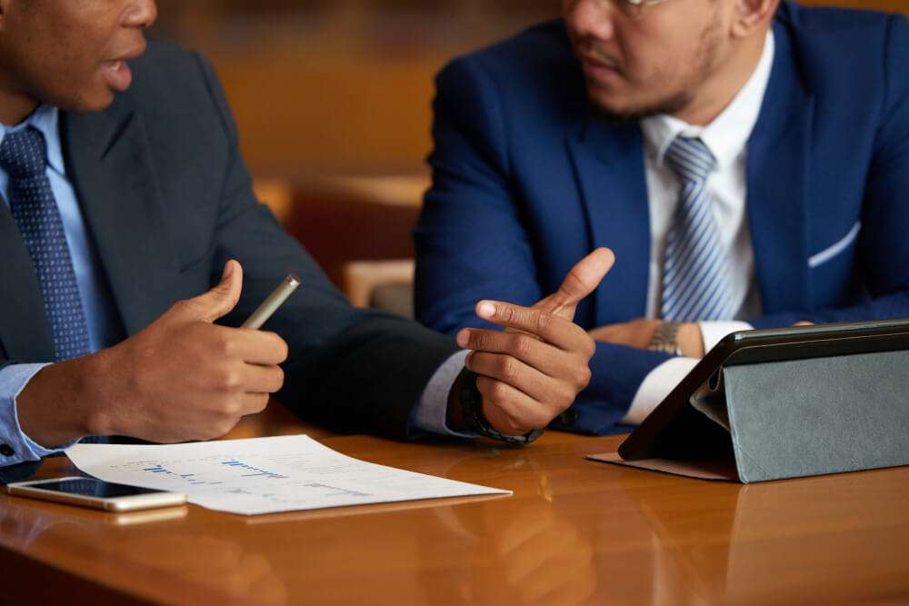 Two businessmen in suits discuss graphs with a tablet and papers at a meeting table.
