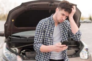 Man with phone puzzled by car breakdown on road, hood open.