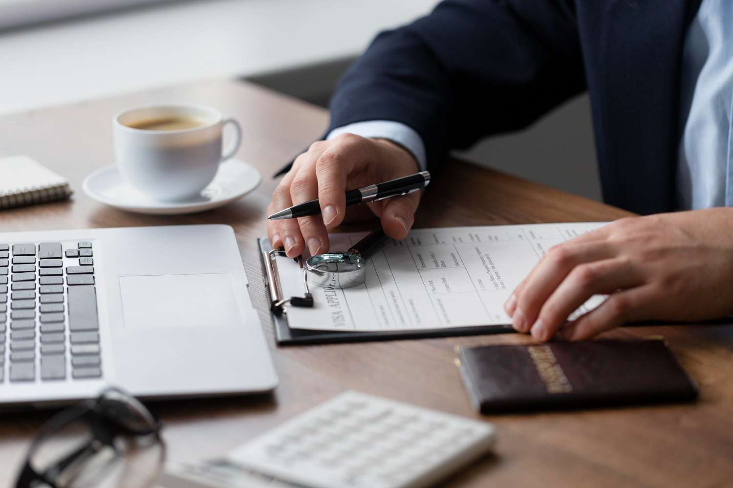 Person filling form on clipboard at desk with laptop and coffee