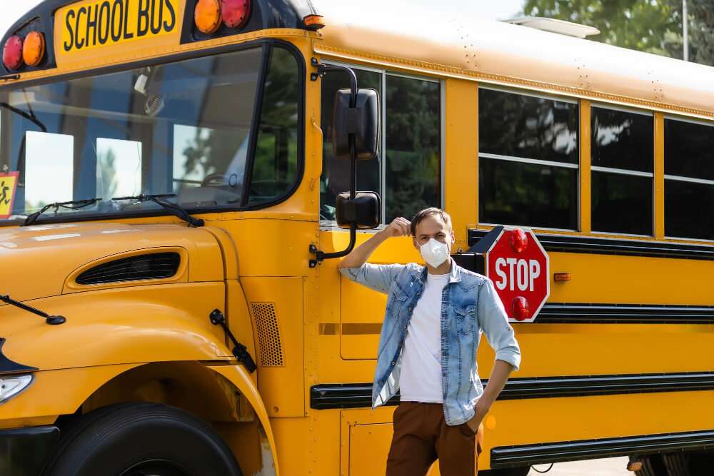 Person in mask stands beside yellow school bus with stop sign.
