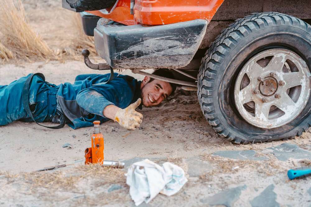 Mechanic giving thumbs up under car with flat tire and jack in desert terrain.