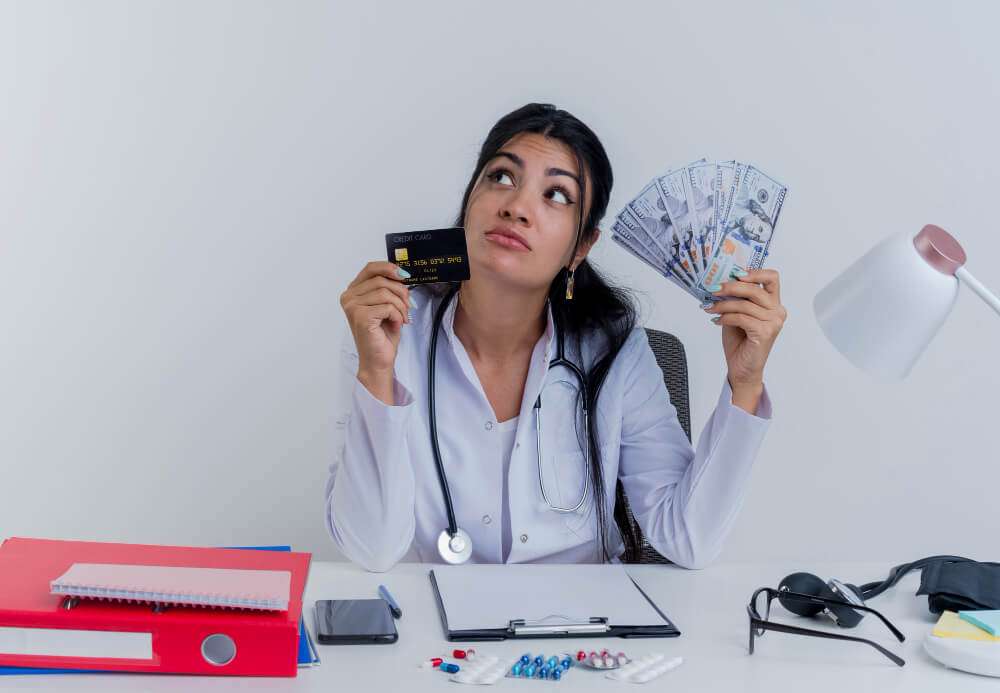 Doctor pondering over credit card and cash at desk with medical tools.