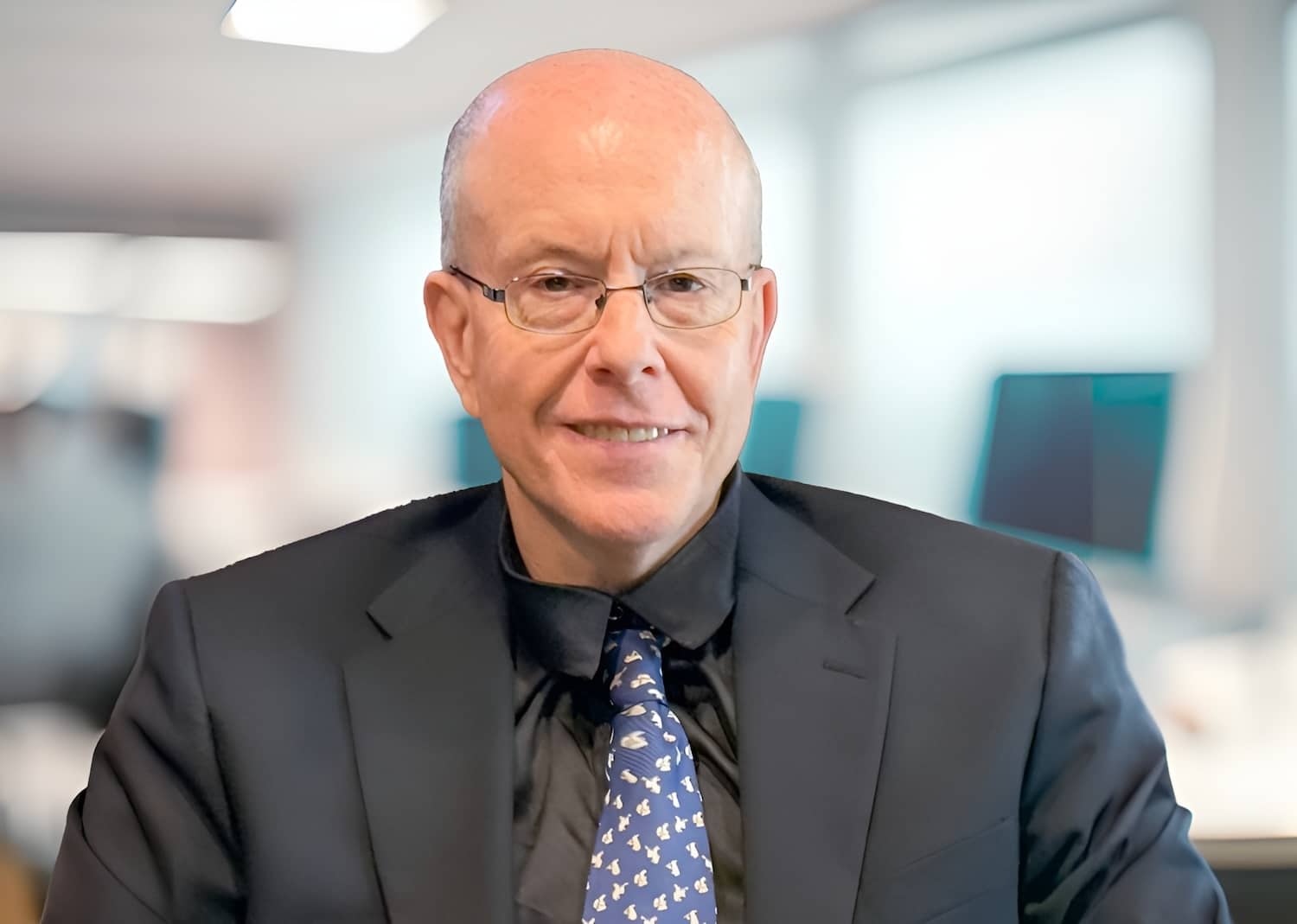 Bald man in dark suit and glasses smiling in an office setting with blurred background.