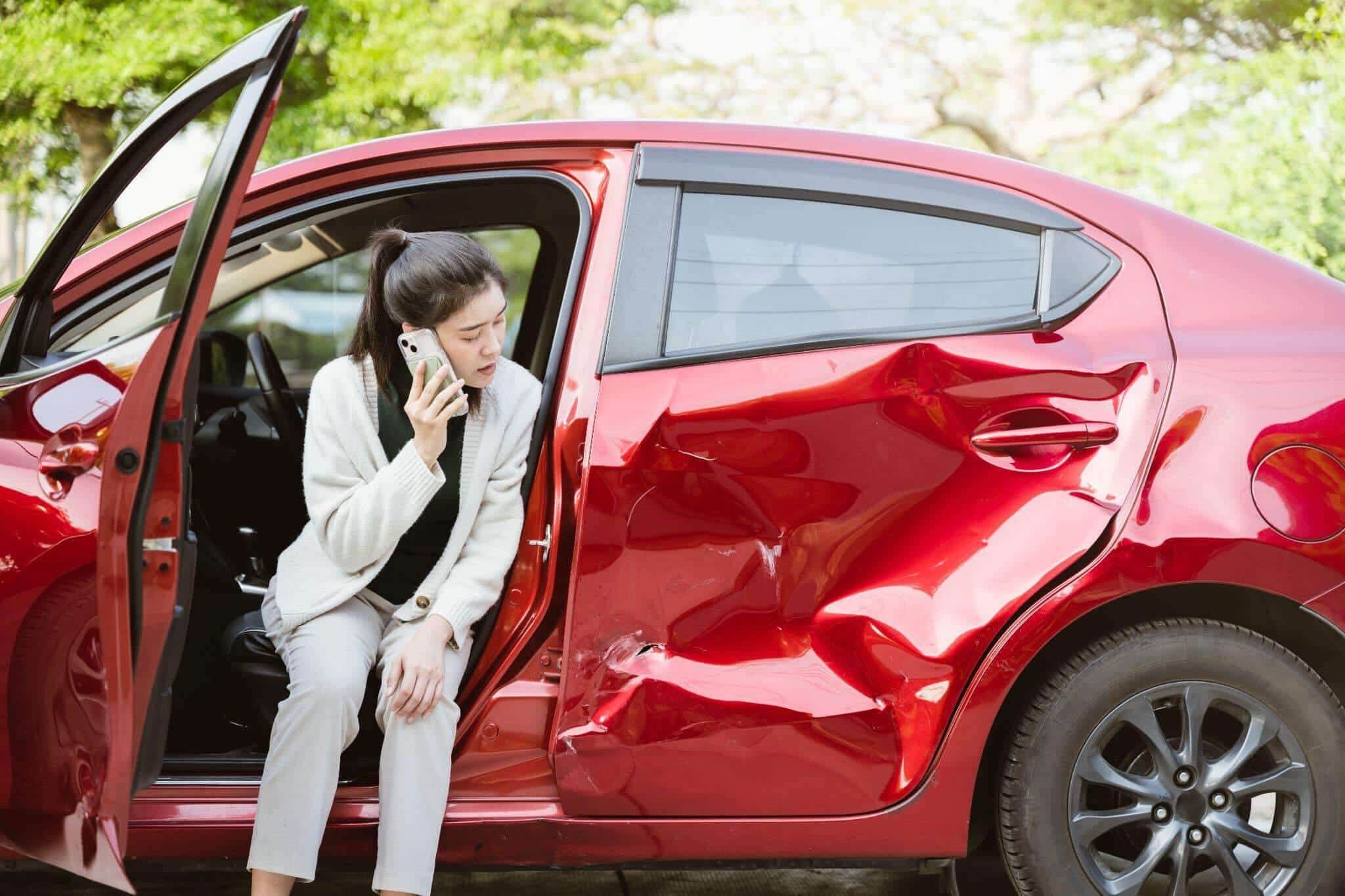 a woman sitting in a red car talking on a cell phone, the car is visibly damaged