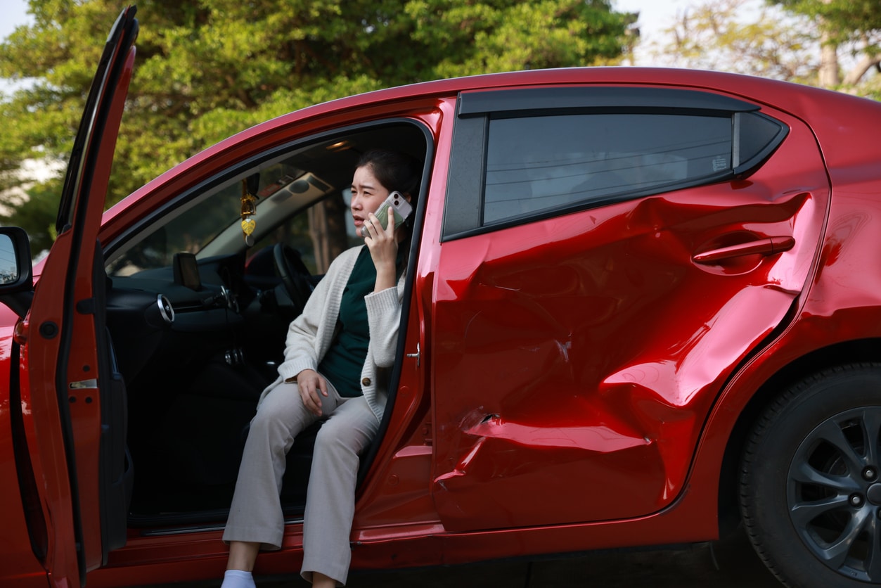 A female motorist is standing on the side of the road after a car accident, assessing the damage to her vehicle. She confidently calls for roadside assistance, detailing her location and the nature of the incident.